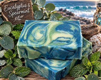 May include: A rectangular bar of soap with a marbled blue and green swirl pattern. A wooden sign with the text Eucalyptus & Spearmint is positioned behind the soap, surrounded by fresh green eucalyptus leaves and sprigs of mint foliage.