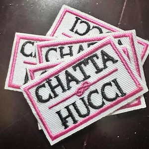May include: A stack of white rectangular fabric patches with pink borders. The patches feature the text "CHATTA & HUCCI" in black, with a pink line separating the words. The patches are stacked on a dark surface.