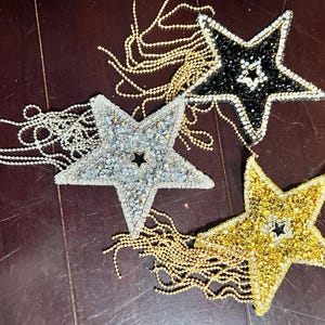 May include: Three star-shaped ornaments with beaded embellishments and dangling chains. One star is silver, one is black, and one is gold. Each star has a smaller black star in the center. The ornaments are on a dark brown surface.