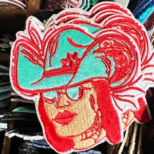 May include: Embroidered patch featuring a woman wearing a turquoise hat with red accents and red hair. The patch has a white border and depicts the woman's face with glasses and red lips. The design is detailed and colorful.