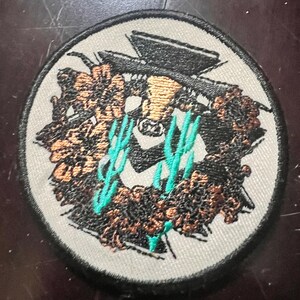 May include: Round embroidered patch with a black border. The design features a stylized skull wearing a hat, surrounded by a wreath of brown and gold elements, with teal accents. The patch is on a light gray background.