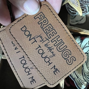 May include: Two brown leather tags with the text "FREE HUGS just kidding DON'T TOUCH ME" in black. The tags are rectangular with rounded corners and stitched edges. The tags are being held by a person.