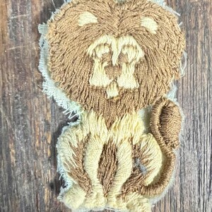 May include: Embroidered lion patch in shades of brown and cream. The lion has a detailed mane, face, and body, with a small tail. The patch is made of textured fabric and is likely designed to be sewn onto clothing or accessories.