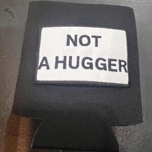 May include: A black foam beverage insulator featuring a rectangular white patch centered on the front. The patch has a black embroidered border and displays the text NOT A HUGGER in bold black capital letters, centered on the white background.