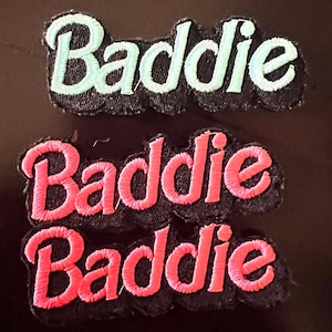 May include: Two embroidered patches with the word "Baddie". One patch features the word in light green thread, and the other two patches have the word in bright pink thread. The patches are on a black background.