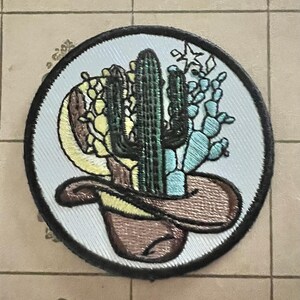 May include: Embroidered patch featuring a cowboy hat with a cactus and other desert plants. The patch has a light blue background and a black border. The cactus is green, and the other plants are yellow and blue.