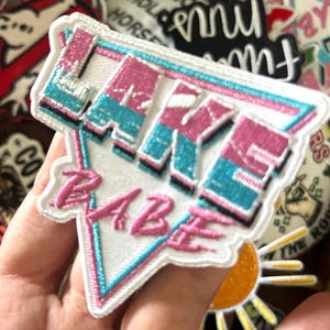 May include: Embroidered patch with the words "Lake Babe" in pink and teal, outlined in white. The patch is in a triangular shape with a retro design. Other patches are visible in the background.