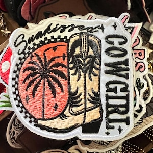 May include: Embroidered patch with a cowboy boot, palm tree, and sunset design. The patch has the words "SunKissed" and "Cowgirl" in a white, stylized font. The patch is white with black, orange, and tan details.
