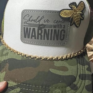 Puede incluir: Un gorro de camionero blanco y camuflaje con un ribete de cadena dorada. El gorro tiene un parche gris con el texto "Should've come with a WARNING" y una aplicación de abeja dorada.