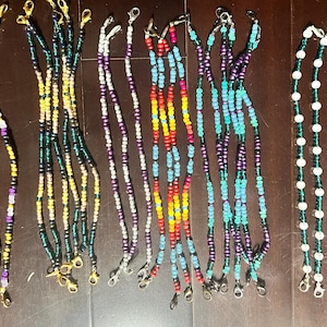 May include: A collection of beaded mask lanyards in various colors. The lanyards feature gold, silver, and black clasps. Colors include purple, yellow, green, blue, red, and white. The beads are strung on a thin cord.