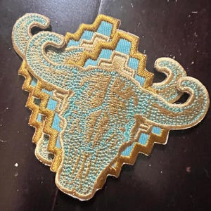 May include: Embroidered patch featuring a stylized bull skull design. The patch has a turquoise and gold color scheme with intricate stitching. The design includes geometric patterns and curved horns, suitable for jackets, bags, or other fabric items.