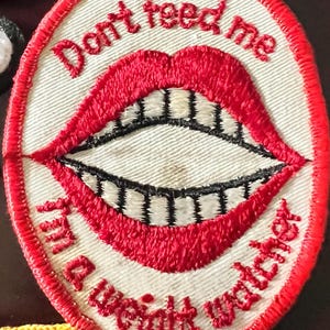 May include: Embroidered patch with a red border and the words "Don't feed me I'm a weight watcher" surrounding a red mouth with black teeth. The patch is white with red stitching.
