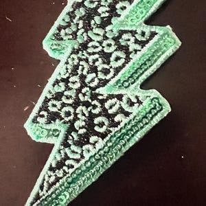 May include: A green and black lightning bolt-shaped patch. The lightning bolt has a green border with a detailed pattern. The interior is black with a white spotted pattern. The patch is likely for clothing or accessories.