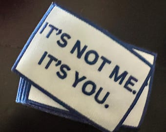 May include: A stack of white rectangular patches with blue stitched borders. The patches have the text "IT'S NOT ME. IT'S YOU." embroidered in blue thread. The patches are stacked on a dark surface.