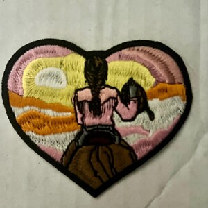 May include: Heart-shaped embroidered patch featuring a cowgirl riding a horse, set against a sunset backdrop. The design includes a pink shirt, a braided hairstyle, and a black border. The patch is suitable for jackets, bags, or other fabric items.