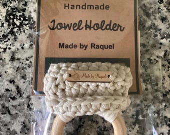 May include: Handmade towel holder made by Raquel. The holder is made of crocheted cream-colored cotton and a natural wood ring. The tag says "Handmade Towel Holder Made by Raquel".