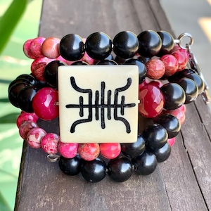May include: A multi-strand bracelet with black and pink beads. The center of the bracelet features a square, ivory-colored charm with a black symbol.