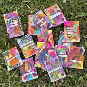 May include: A collection of colorful, abstract art cards with various text snippets. The cards feature mixed media designs with vibrant hues of pink, orange, blue, and green. The cards are scattered on a bed of green grass.