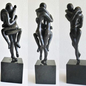 Puede incluir: Una escultura de bronce de dos figuras abrazándose, la figura más pequeña es sostenida en alto por la figura más grande. La escultura está montada sobre una base cuadrada negra.