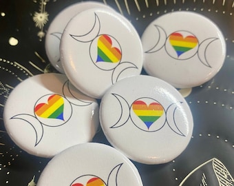 May include: Five white button pins with a rainbow heart inside a triple moon symbol. The pins are on a black background with a white star and a circle design.