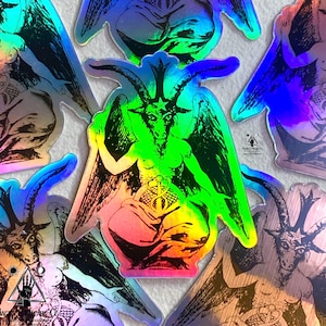 May include: A set of five iridescent holographic stickers featuring a black and white illustration of a winged, horned figure with a pentagram on its forehead. The figure is sitting with its legs crossed and holding a snake in its hands. The stickers are all different sizes and shapes, and they are all very colorful.