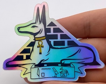 Puede incluir: Una pegatina holográfica con un chacal Anubis con un ankh dorado, sentado en una pirámide con un patrón de ladrillos en blanco y negro. La pegatina también incluye un pájaro pequeño y un símbolo jeroglífico.