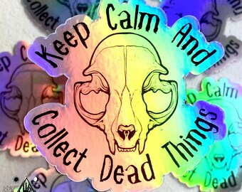 Puede incluir: Una pegatina holográfica con un fondo degradado arcoíris. La pegatina presenta una ilustración en blanco y negro de una calavera de gato con el texto "Keep Calm And Collect Dead Things".