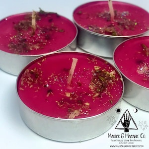 May include: Four pink tea light candles with a gold glitter and dried herbs embedded in the wax. The candles are in silver holders. The text on the label reads "Magick & Macabre Co. Pagan Supplies, Vegan Bath Products, & Cosmic Goods www.magickandmacabre.co.com"
