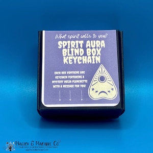 May include: A small black box with a purple and white label that says "What spirit calls to you? Spirit Aura Blind Box Keychain". The label also features a white and yellow Ouija planchette with a message for you.