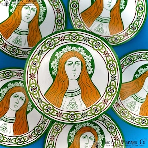 May include: A round sticker with a green, gold, and white Celtic knot border. The center features a woman with long orange hair wearing a white dress and a green wreath. She has a green Celtic knot on her chest. The text "MAGICK & MACABRE CO." is at the bottom of the sticker.