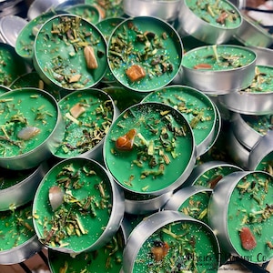 Puede incluir: Una colección de velas de té verdes en recipientes de metal plateado. Cada vela está adornada con hierbas secas y una pequeña piedra preciosa. Las velas se muestran en primer plano.