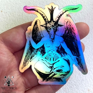 May include: A holographic sticker featuring a black and white illustration of a goat-headed figure with wings, sitting in a meditative pose. The figure has a star on its head and a snake wrapped around its waist. The sticker has a rainbow iridescent finish.