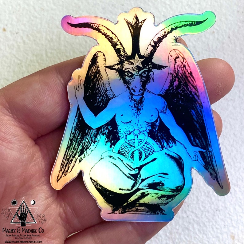 May include: A holographic sticker featuring a black and white illustration of a goat-headed figure with wings, sitting in a meditative pose. The figure has a star on its head and a snake wrapped around its waist. The sticker has a rainbow iridescent finish.