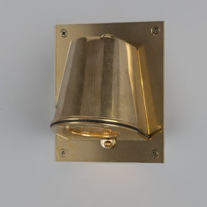 Puede incluir: Una lámpara de pared de color dorado con una pantalla redonda en forma de campana. La lámpara tiene un diseño simple y moderno y está montada sobre una base cuadrada.