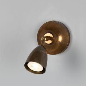 Op de afbeelding: Een wandlamp met een ronde achterplaat en een conische lampkop, beide uitgevoerd in een geborstelde antiek messing afwerking. Een verstelbare arm verbindt de basis met de lamp die een zacht, gericht licht uitstraalt.