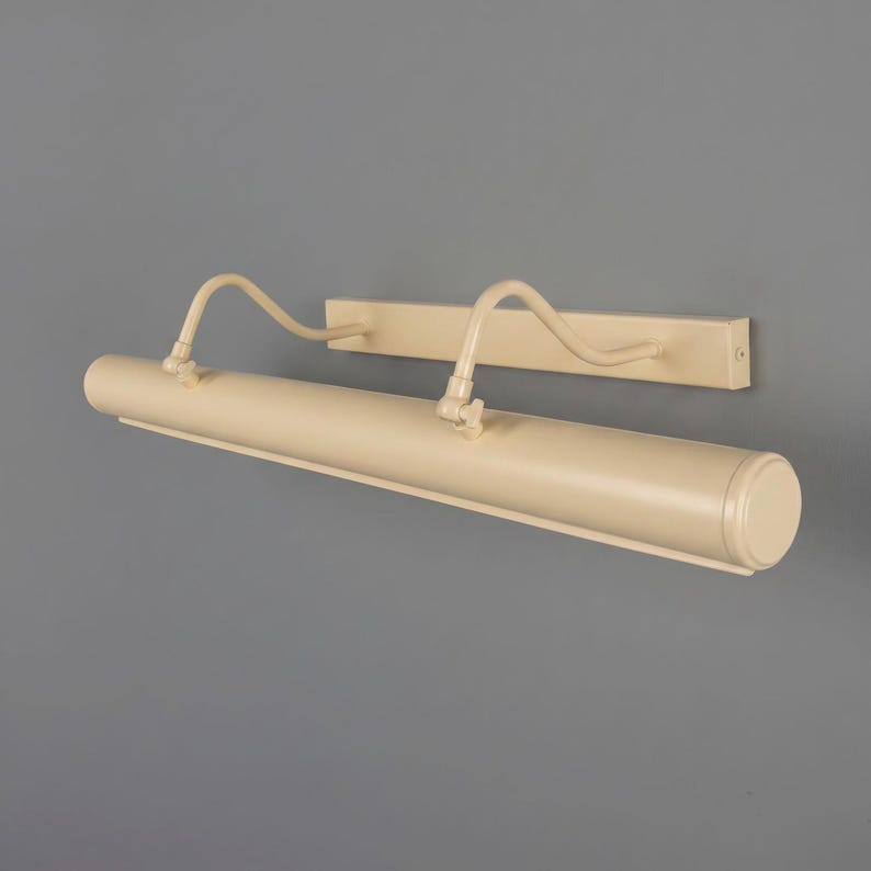 May include: A cream-colored metal picture light fixture featuring a long cylindrical lamp head attached to a rectangular wall-mounting plate by two curved, adjustable arms with visible hinge joints for directing the light beam.