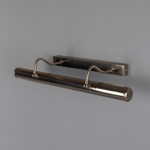 May include: A wall-mounted picture light with a metallic finish. The fixture features a long, cylindrical horizontal lamp head attached to a rectangular backplate by two curved, adjustable arms that extend outward from the wall mount.
