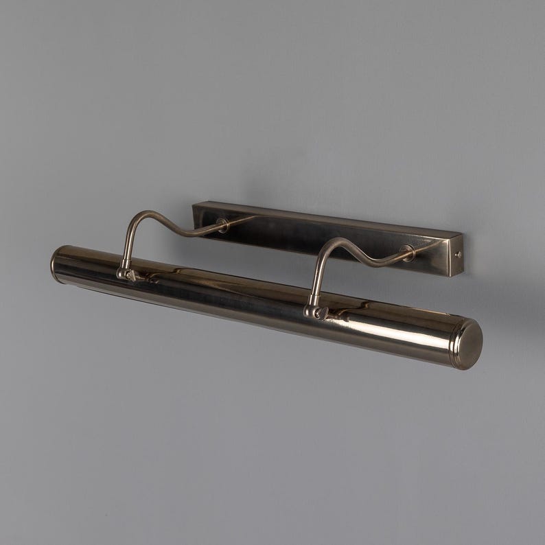 May include: A wall-mounted picture light with a metallic finish. The fixture features a long, cylindrical horizontal lamp head attached to a rectangular backplate by two curved, adjustable arms that extend outward from the wall mount.