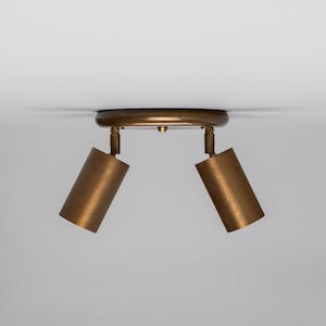 Puede incluir: Un foco de techo de dos luces de color bronce. La luminaria tiene una base circular y dos focos cilíndricos que se pueden ajustar. Los focos están orientados hacia abajo.