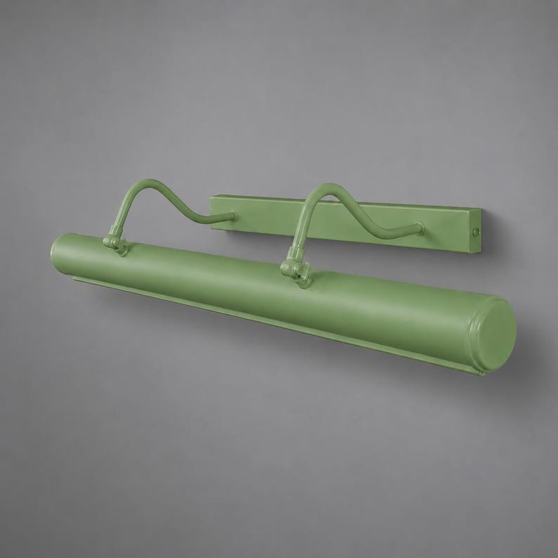 May include: A sage green picture light fixture featuring a long, cylindrical lamp head attached to a rectangular wall-mounting plate by two curved, adjustable metal arms. The entire unit has a smooth, matte finish and a minimalist, functional design.