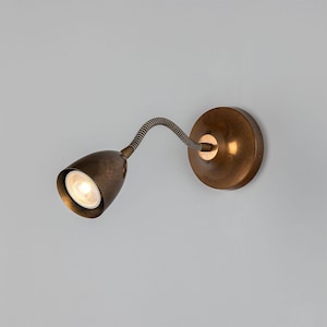 May include: A wall-mounted lamp with an antique brass finish, featuring a circular base, a flexible gooseneck arm, and a conical spotlight head. The lamp includes a single integrated light bulb that emits a warm glow from the centre.