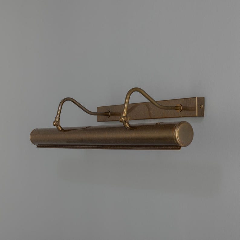 May include: A wall-mounted picture light with a metallic bronze finish. The fixture features a rectangular backplate, two curved support arms, and a cylindrical light housing that extends horizontally across the front of the unit.
