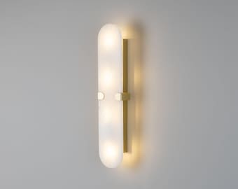 May include: A vertical wall sconce featuring a long, rounded, translucent white glass panel illuminated from within. The glass is secured to a rectangular gold-toned metal base by two small, matching gold-toned metal clips on the sides.