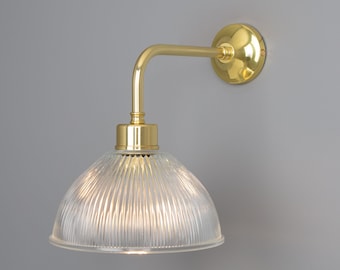 May include: A wall-mounted light fixture featuring a polished brass arm and a circular wall plate. The lamp includes a clear, ribbed glass dome-shaped shade that directs light downward, connected to the brass hardware by a cylindrical socket.