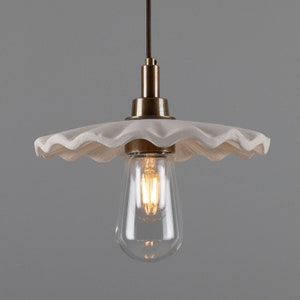 May include: White ceramic pendant light fixture with a scalloped edge and a brass finish. The light fixture is hanging from a black cord and has a clear glass bulb.