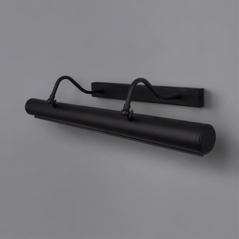 May include: A matte black picture light fixture featuring a long cylindrical lamp head attached to a rectangular wall-mounting plate by two curved, adjustable metal arms. The design includes visible pivot joints for angling the light source.