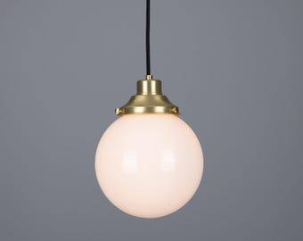 May include: A spherical pendant light fixture featuring a smooth, opaque white glass globe shade attached to a brushed brass metal cap. The light hangs from a thin black cord, providing a clean, balanced appearance for interior lighting.