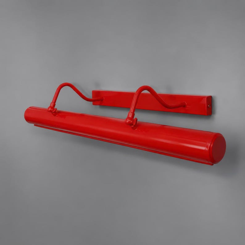 May include: A bright red wall-mounted picture light featuring a long cylindrical lamp head attached to a rectangular backplate by two curved, adjustable metal arms. The entire fixture has a smooth, glossy finish and a minimalist, linear design.