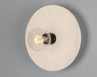 May include: A round, off-white wall sconce with a textured, sunburst-like design. It features a clear glass bulb and a black base. The sconce is mounted on a gray wall, providing a neutral backdrop for the fixture.