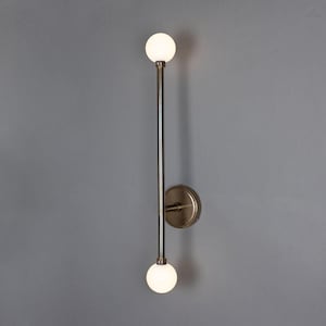 May include: A modern wall sconce with a brushed brass finish and two white globe light bulbs. The sconce has a long, vertical arm with the bulbs positioned at the top and bottom.